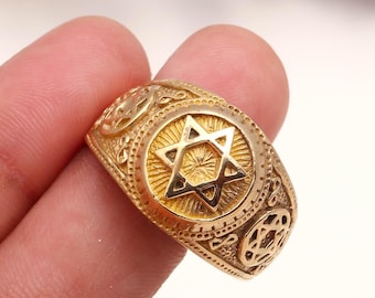 May include: A gold-colored ring featuring a Star of David in the center, surrounded by a circular design with radiating lines. The ring has intricate patterns and symbols along the band, with a polished finish. The ring is held by a person's fingers.