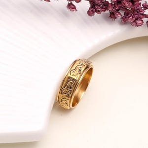 Può includere: Un anello color oro con un disegno inciso di cavalli. L'anello è posto su uno sfondo bianco con un rametto di fiori viola nell'angolo in alto a destra. L'anello è uno stile a fascia.