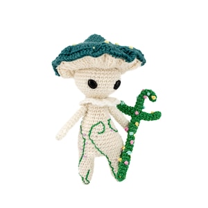 Peut inclure: Un personnage de champignon crocheté avec un chapeau turquoise et un corps blanc, tenant un bâton vert avec des points colorés. Le personnage a des yeux noirs et une expression fantaisiste.