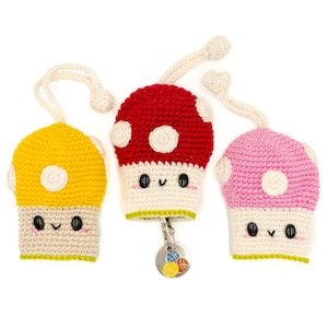 May include: Three crocheted mushroom keyrings with smiling faces. The mushrooms are red, yellow, and pink with white spots. Each keyring has a green loop and a silver key ring.