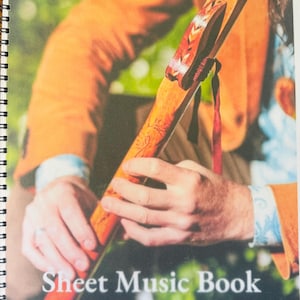 Può includere: Un libro di spartiti intitolato "Aris Flutes" con il testo "Sheet Music Book 2023". La copertina presenta una persona che tiene un flauto di legno. L'indirizzo del sito web www.arisflute.com è visibile in basso.