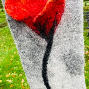May include: A grey wool scarf with a red felt poppy flower appliqué. The flower has a black stem and is centred on the scarf.