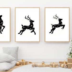 May include: Three framed black and white silhouette prints of reindeer with a sparkling effect. The reindeer are in different poses, one is standing, one is running, and one is jumping. The prints are displayed on a white wall with a Christmas tree and gifts in the foreground.