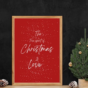 May include: A red framed print with white text that reads "The true spirit of Christmas is Love." The background of the print is red with a white snow effect. A small Christmas tree with gold baubles is in the foreground.