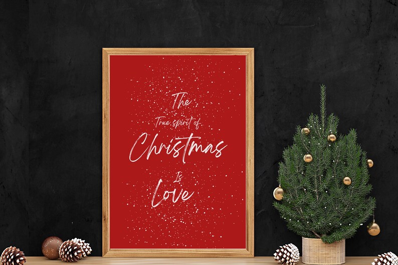 May include: A red framed print with white text that reads "The true spirit of Christmas is Love." The background of the print is red with a white snow effect. A small Christmas tree with gold baubles is in the foreground.