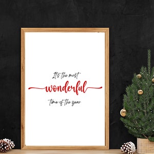 May include: A framed print with the text "It's the most wonderful time of the year" in red cursive lettering on a white background. The frame is brown wood and the print is sitting on a wooden surface in front of a black wall. A small potted Christmas tree with gold baubles is to the right of the print.