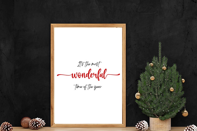 May include: A framed print with the text "It's the most wonderful time of the year" in red cursive lettering on a white background. The frame is brown wood and the print is sitting on a wooden surface in front of a black wall. A small potted Christmas tree with gold baubles is to the right of the print.