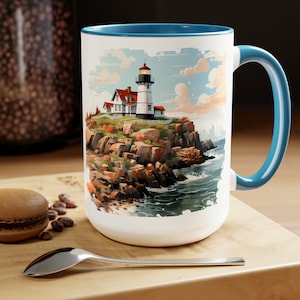 May include: White ceramic mug with a blue rim and handle. The mug features a colorful illustration of a lighthouse on a rocky coastline with ocean waves.