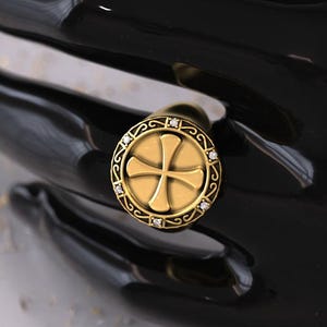 Könnte beinhalten: Ein goldener Ring mit einem Kreuzdesign in einem kreisförmigen Rahmen, verziert mit kleinen, klaren Edelsteinen. Das Band des Rings ist schlichtes, poliertes Gold. Der Ring wird auf einer schwarzen Oberfläche gezeigt.