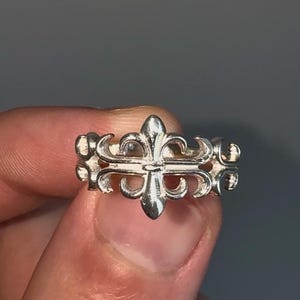 May include: A silver ring featuring a fleur-de-lis design. The ring has a repeating pattern of the stylized floral emblem, with a polished finish. The ring is held in a hand against a gray background.