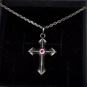 May include: A silver cross pendant necklace with a pink gemstone center, displayed in a black jewelry box. The cross has pointed ends and is suspended from a delicate chain. The box has a silver logo on the lid.