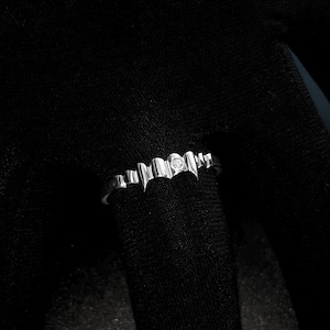 May include: A silver ring with a repeating heart-shaped design and a small, round diamond. The ring is set against a dark background, highlighting its delicate details and shine. A piece of jewelry.