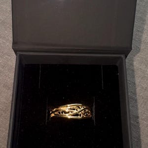May include: A gold ring with a delicate, intricate design sits in a black velvet ring box. The box is closed and has the letters "PLI" embossed on the top.