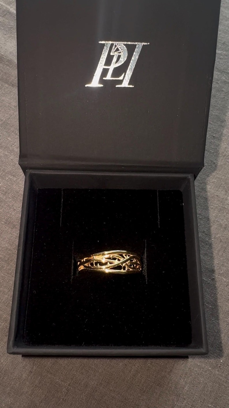 May include: A gold ring with a delicate, intricate design sits in a black velvet ring box. The box is closed and has the letters "PLI" embossed on the top.
