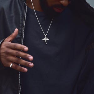 May include: A silver pendant necklace featuring a detailed angel design. The necklace is worn over a black shirt and is paired with a black leather jacket. A silver ring is visible on the hand.