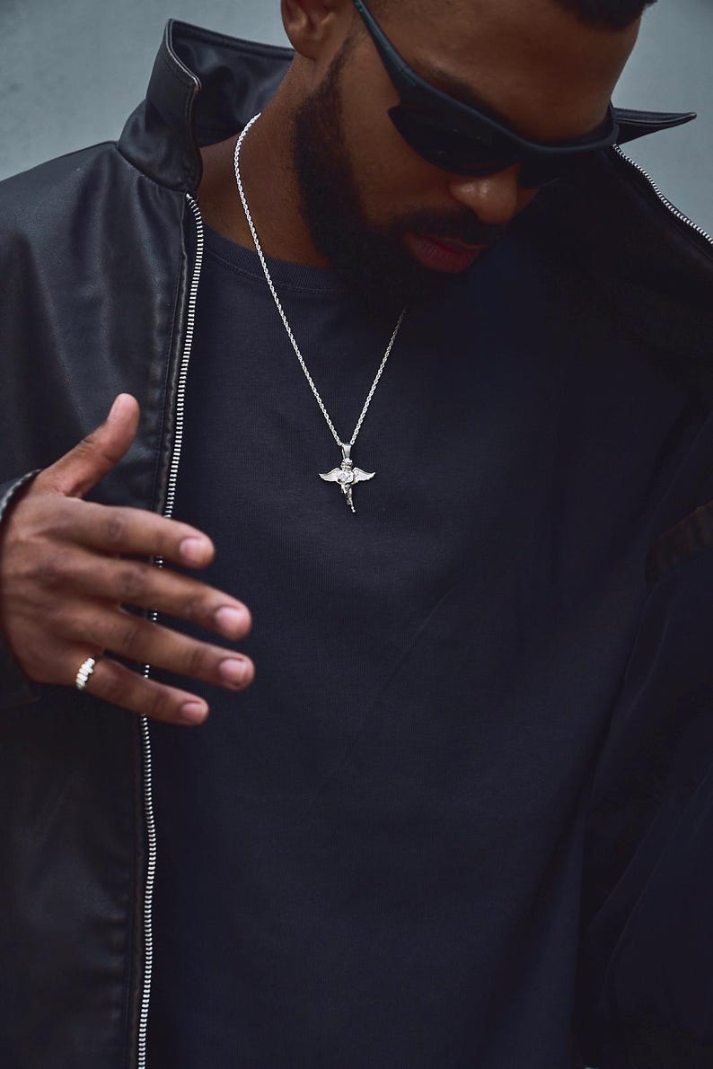 May include: A silver pendant necklace featuring a detailed angel design. The necklace is worn over a black shirt and is paired with a black leather jacket. A silver ring is visible on the hand.
