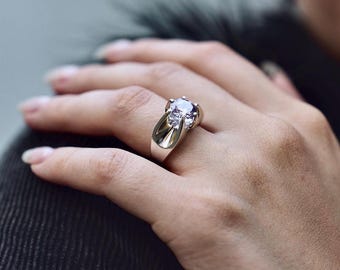 Puede incluir: Un anillo de plata con una gran piedra preciosa redonda y transparente engastada en garras. El anillo tiene un diseño único y curvo.