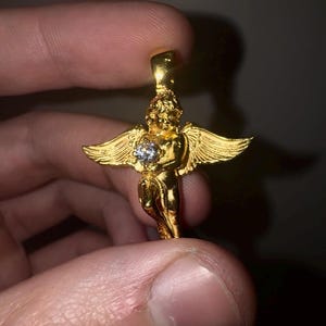 May include: A gold-colored angel pendant with outstretched wings and a central, clear gemstone. The pendant is held between fingers, showcasing its intricate details and reflective surface. The angel figure is a classic jewelry design.