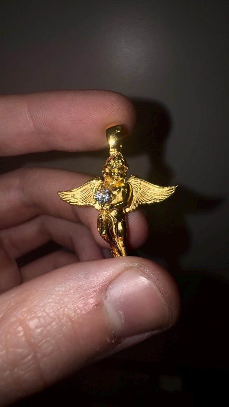 May include: A gold-colored angel pendant with outstretched wings and a central, clear gemstone. The pendant is held between fingers, showcasing its intricate details and reflective surface. The angel figure is a classic jewelry design.