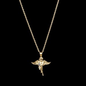 May include: A gold-colored angel pendant necklace. The pendant features a detailed cherub with outstretched wings, holding a small gem. The necklace chain is also gold and hangs against a black background.