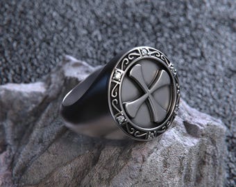 May include: A silver signet ring with a cross design. The ring features a circular face with a cross motif, surrounded by a decorative border with small, clear stones. The band is smooth and polished.