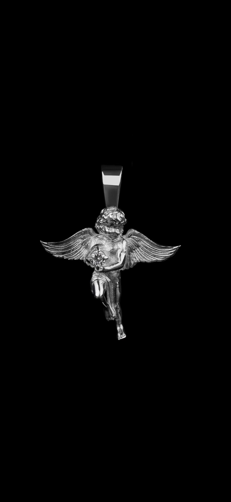 May include: Silver pendant featuring a detailed cherub with outstretched wings, holding a gem. The pendant has a polished finish and is suspended from a rectangular bail. The background is solid black, highlighting the pendant's design.