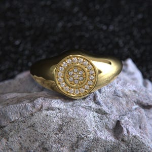 May include: A gold ring with a circular design of small, clear gemstones. The ring is set on a gray stone surface against a dark background. The ring's band is smooth and rounded.