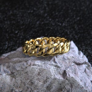 May include: A gold-colored chain ring with a curb link design. The ring is displayed on a gray rock against a dark background. The ring's links are closely interlocked, creating a continuous, textured surface. The ring is a unisex jewelry item.
