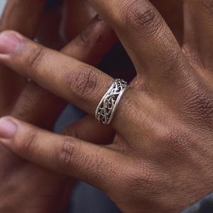 May include: A silver ring with a detailed, intricate design. The ring is worn on a person's finger.
