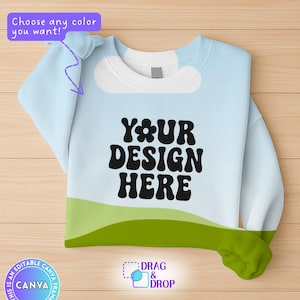 Puede incluir: Una sudadera de color azul claro con un diseño de paisaje verde y blanco. El texto "YOUR DESIGN HERE" está impreso en negro en la parte delantera de la sudadera.