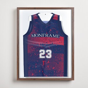 May include: Framed navy blue basketball jersey with red accents and the word "MONFRAME" across the chest. The number "23" is displayed in white. The jersey is mounted on a white hanger inside a brown wooden frame.