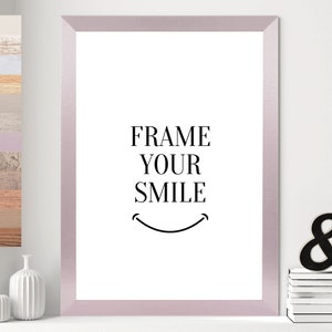Op de afbeelding: Een wit ingelijst print met de tekst "FRAME YOUR SMILE" in zwarte letters. Onder de tekst staat een simpel zwart-wit smiley-gezicht.