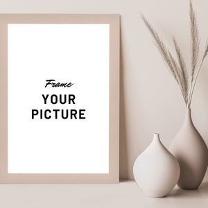 May include: A beige picture frame with a white mat and the text "Frame Your Picture" inside. The frame is sitting on a white surface next to two beige ceramic vases with dried pampas grass.