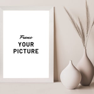 Può includere: Una cornice per foto bianca con un passepartout bianco e uno sfondo bianco. La cornice è appoggiata su una superficie marrone chiaro. Il testo "Frame Your Picture" è scritto in nero all'interno della cornice.