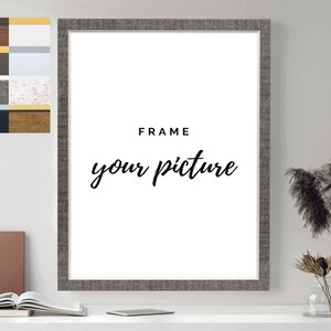 Op de afbeelding: Een grijs fotolijstje met een witte passe-partout en de tekst "FRAME your picture" op een witte achtergrond. Het lijstje staat op een wit oppervlak met een boek en een telefoon.