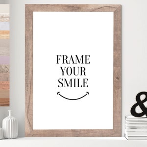 Op de afbeelding: Een ingelijst print met de tekst "FRAME YOUR SMILE" en een simpele zwarte smiley. De lijst is van lichtbruin hout.