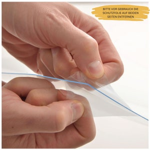 May include: A hand peeling off a clear protective film from a surface. The film has a blue line running along the edge. The text "Bitte vor Gebrauch die Schutzfolie auf beiden Seiten entfernen" is visible in the background.