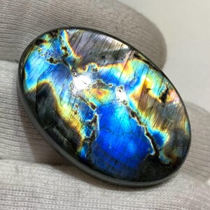 May include: An oval labradorite gemstone with a polished surface. The stone exhibits a vibrant play of colour, with iridescent blues, yellows, and greens. The gemstone has a dark grey border and is set against a white background.