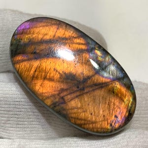 Könnte beinhalten: Ein ovaler Labradorit-Edelstein mit polierter Oberfläche. Der Stein zeigt ein lebendiges Farbspiel mit Orange-, Lila- und Blautönen. Die Oberfläche des Edelsteins hat eine reflektierende Qualität, die seine visuelle Attraktivität erhöht.