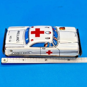 May include: Vintage white toy ambulance with red crosses and the word "AMBULANCE" printed on the side. The toy car has blue window details and a driver figure. The number "19" is printed on the side.