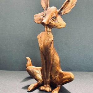 May include: A bronze-toned sculpture of a stylized fox sitting in an upright position. The figure features elongated ears, a slender neck, and a textured surface with visible brushstrokes. The fox has a pointed snout and dark, almond-shaped eyes.