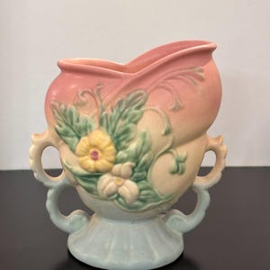 May include: A ceramic vase featuring a pastel color palette of pink, yellow, and light blue. The vessel has two ornate handles on the sides and a raised relief design on the front depicting green leaves and two flowers in yellow and white.