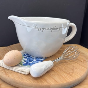 May include: A white ceramic mixing bowl with a pouring spout and handle, inscribed with "happy everything." A whisk and an egg rest on a wooden cutting board. A small floral patterned cloth and a speckled egg are also present.