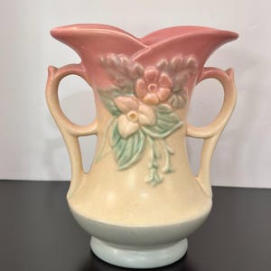 May include: A ceramic vase featuring a two-handled design with a scalloped rim. The piece displays a gradient color scheme transitioning from a soft pink at the top to a cream center and a pale blue base, accented by a raised floral relief.