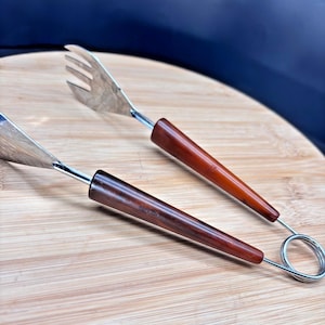 May include: A pair of vintage salad servers with stainless steel heads and dark brown wooden handles. The serving fork has three tines. The servers are resting on a wooden cutting board.