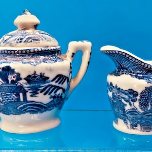 May include: A vintage porcelain sugar bowl with a lid and a creamer. Both pieces feature a blue and white Willow pattern design. The sugar bowl has two handles and a finial on the lid. The creamer has a spout and a handle.