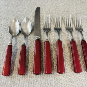 May include: A set of seven pieces of flatware featuring two spoons, one knife, and four forks. Each utensil has a polished silver-tone metal head and a solid, smooth, deep red handle. The items are arranged in a row on a neutral textured surface.