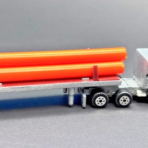 May include: A silver and white toy semi-truck with a red stripe detail, pulling a flatbed trailer loaded with two orange pipes. The trailer has a red deck and silver trim. The truck has six wheels.
