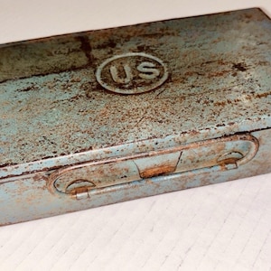 May include: A rectangular, vintage metal box with a faded blue-green paint and significant rust. The lid features a circular emblem with the letters "US". The box has a hinged lid and a latch closure, showing signs of age and wear.