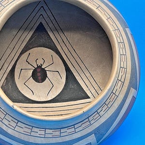 May include: Handmade pottery bowl with geometric designs in black, tan, and gray. The interior features a black triangle and a circle with a spider illustration. The bowl has a wide rim with repeating patterns.